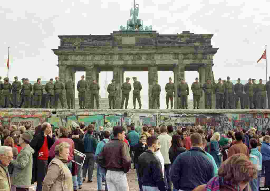 Polițiștii de frontieră din Berlinul de Est stau pe Zidul Berlinului, în fața Porții Brandenburg, pe 11 noiembrie 1989. Cu două zile mai devreme, pe 9 noiembrie, zidul a fost străpuns de mii de germani după ce un oficial est-german a indicat că restricțiile de călătorie între Germania de Est și Germania de Vest urmau să fie eliminate &bdquo;fără întârziere&rdquo;. &nbsp;